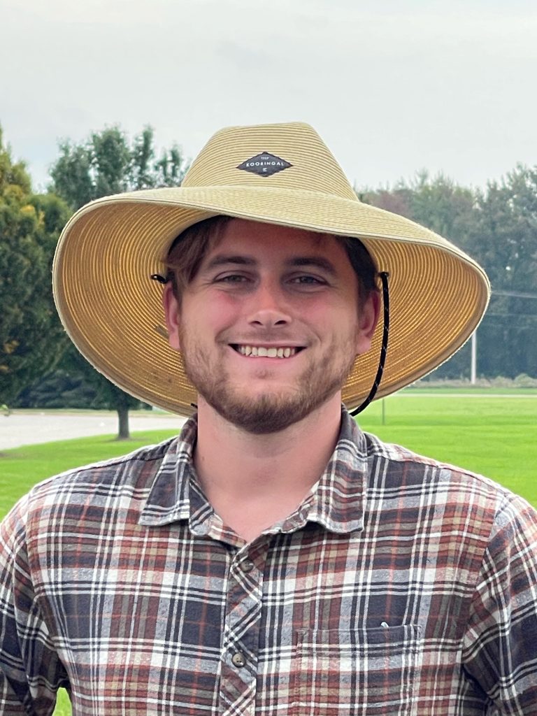 A man wearing a straw hat standing in a field with trees in the background.